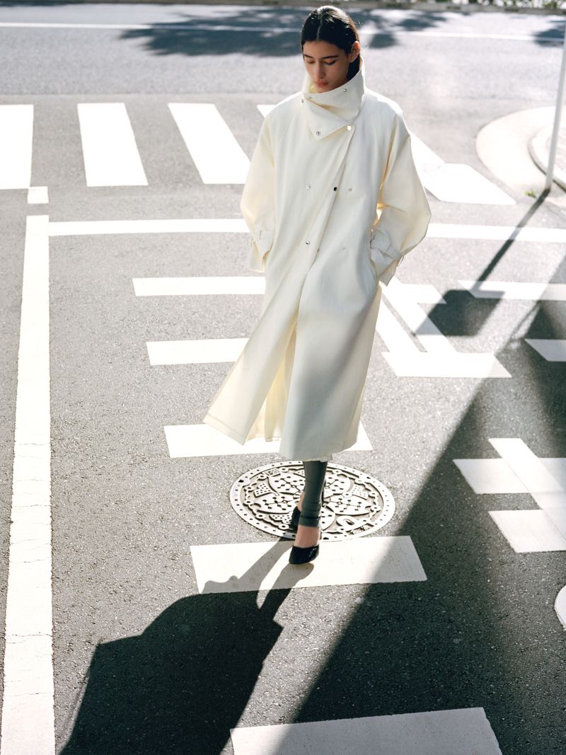 A woman in a long, light coat walks across a crosswalk on a sunny day, with her shadow cast on the grey asphalt.