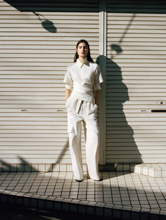 A full-length shot features an adult woman in a white wrapped top and trousers, standing against a closed shutter door outdoors in daylight.