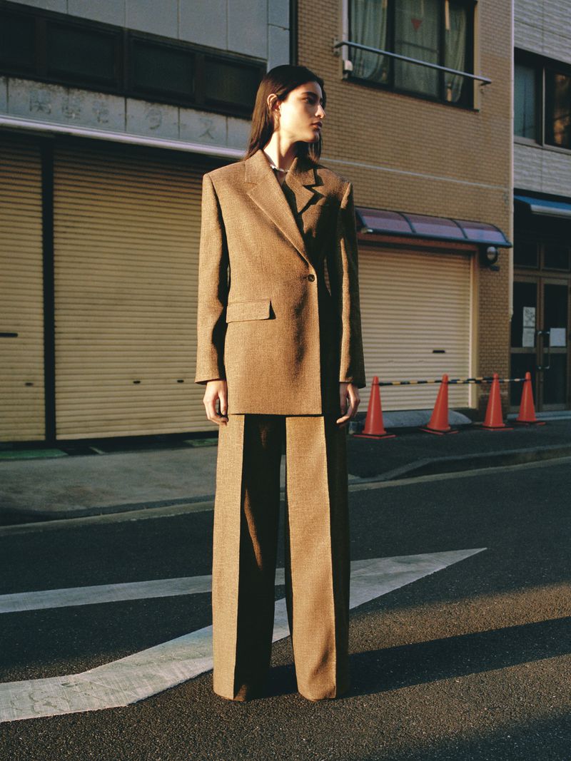 A female fashion model in a brown suit poses on a city street next to a building on an overcast day, showcasing modern fashion.