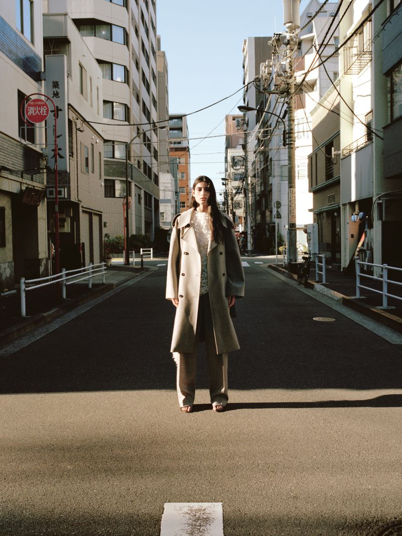 Fashion shot in a city street featuring an adult female model wearing a trench coat and pants, with surrounding buildings and power lines.