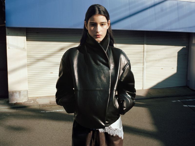 A brunette woman in a black leather jacket poses outdoors on a city street with a garage door and overcast sky in the background.