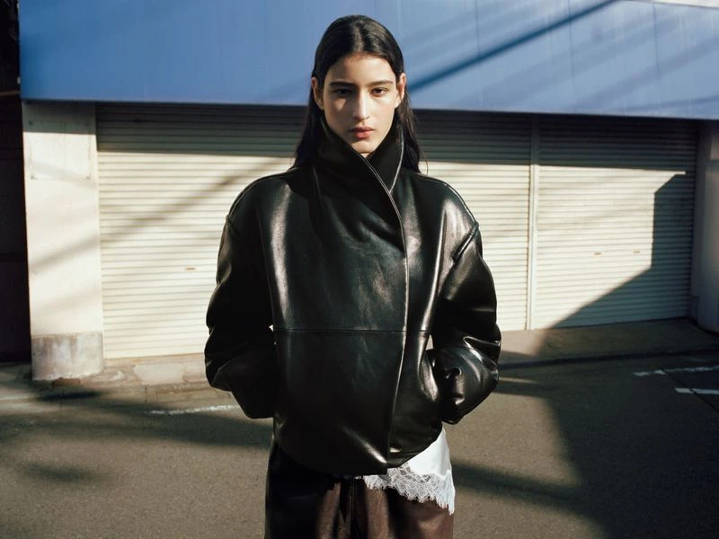 A brunette woman in a black leather jacket poses outdoors on a city street with a garage door and overcast sky in the background.