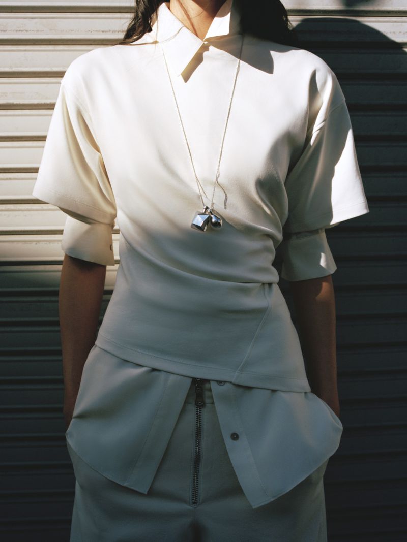 Woman poses wearing a white collared top and matching pants against a corrugated metal background, accessorized with a silver pendant necklace.