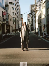 Model standing centrally on a narrow Tokyo street wearing a beige trench coat and trousers, flanked by multi-story buildings.