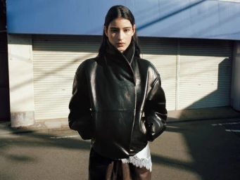 Woman wearing a black leather jacket standing on an urban street in front of a closed storefront shutter.