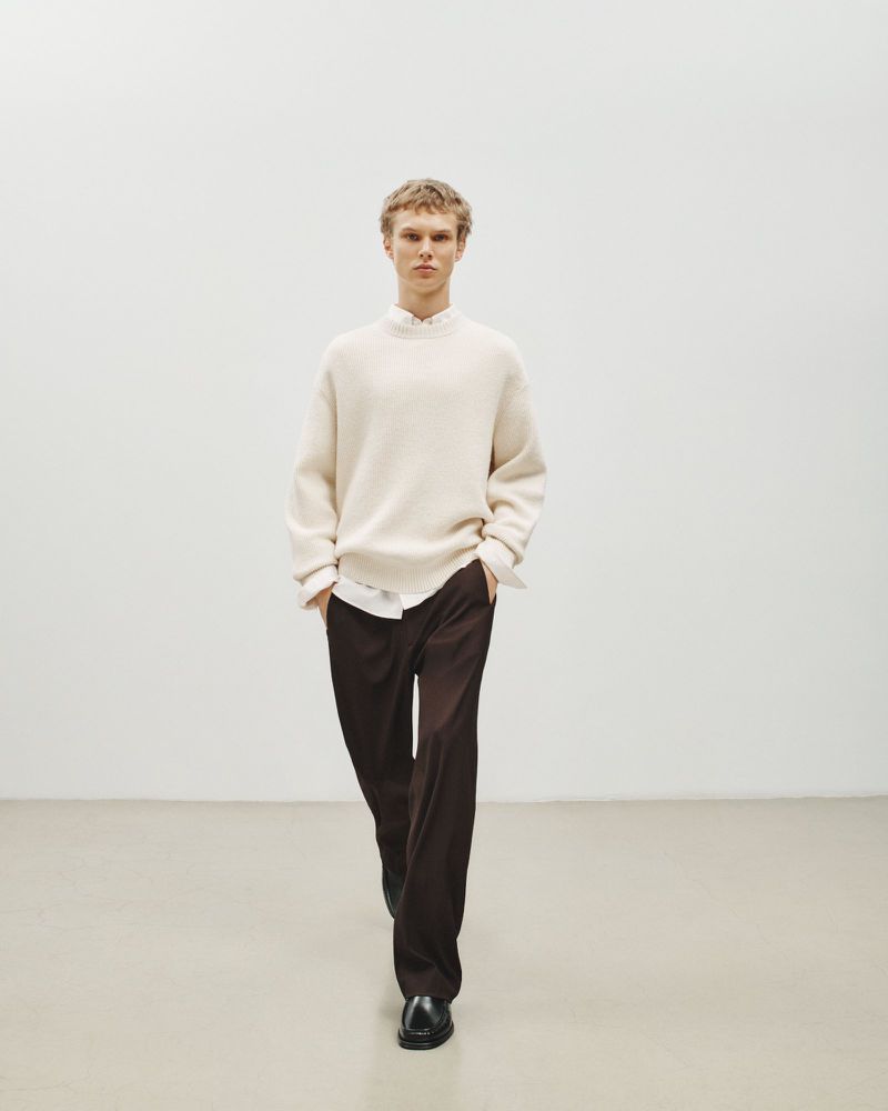 Man in cream sweater and brown pants walking in a minimal, light-filled interior with a neutral color palette. Fashion shot.