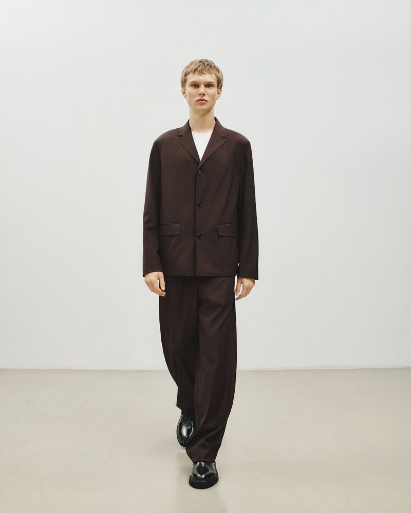 A fair-skinned male model in a brown suit and black dress shoes is walking in a minimalist white studio setting, creating a fashion editorial look.