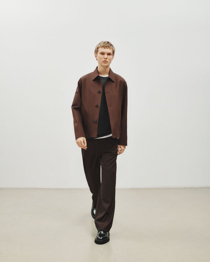 A young adult poses indoors wearing a brown button-down jacket, black shirt, brown trousers, and black loafers against a white backdrop.