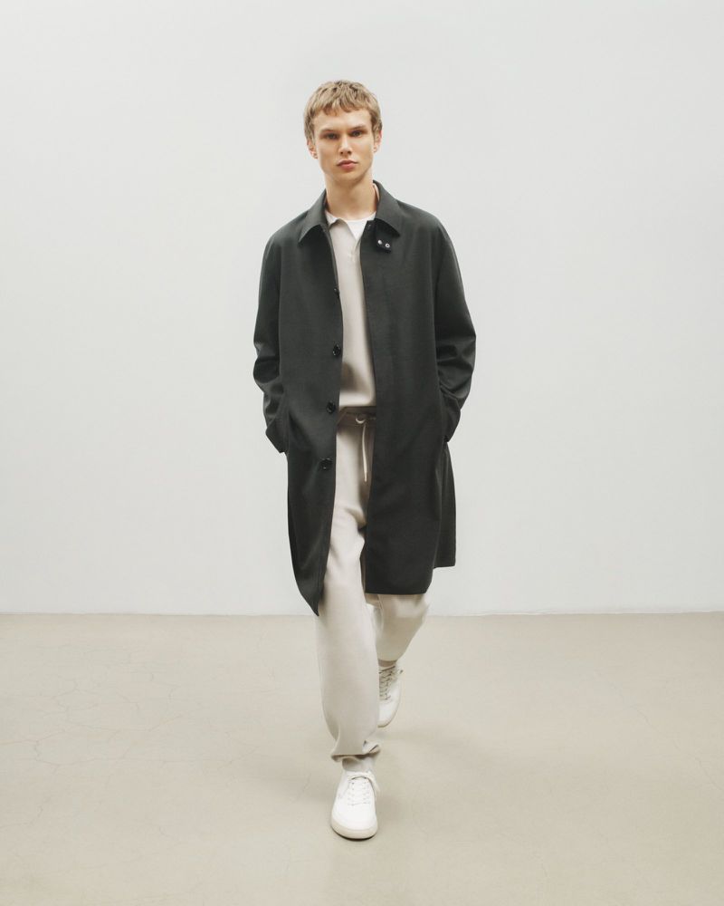 A blond-haired male model is walking indoors wearing a black overcoat, white sweater, white sweatpants, and white sneakers in a fashion editorial.