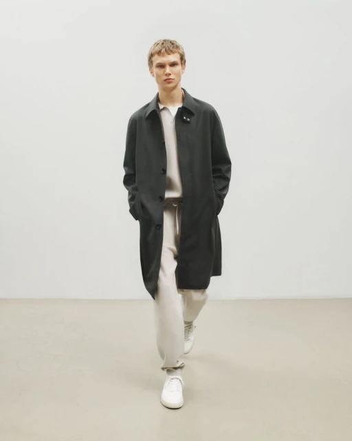 A blond-haired male model is walking indoors wearing a black overcoat, white sweater, white sweatpants, and white sneakers in a fashion editorial.