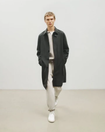 A blond-haired male model is walking indoors wearing a black overcoat, white sweater, white sweatpants, and white sneakers in a fashion editorial.