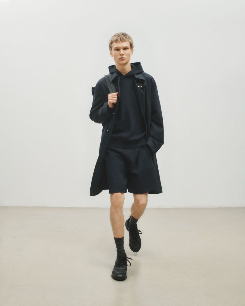 Male model walks in dark hooded sweatshirt, shorts, overcoat, socks, and athletic shoes against a neutral studio backdrop. Fashion shoot.
