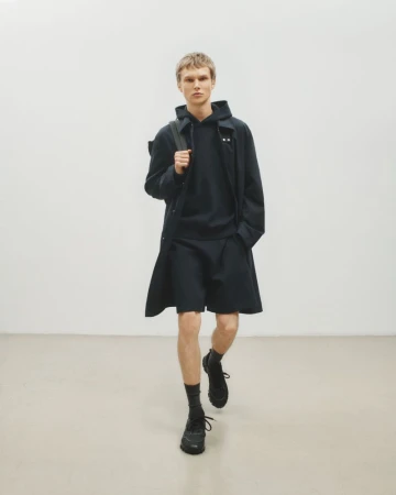 Male model walks in dark hooded sweatshirt, shorts, overcoat, socks, and athletic shoes against a neutral studio backdrop. Fashion shoot.