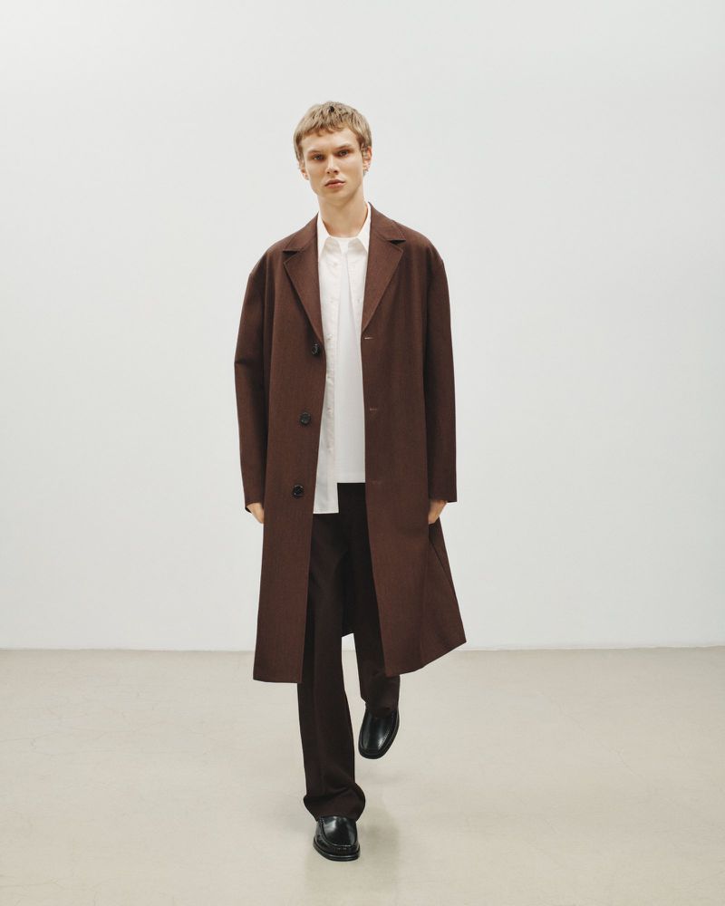 Full-length shot of an adult male wearing a brown coat, white shirt, and brown pants standing in front of a white wall.