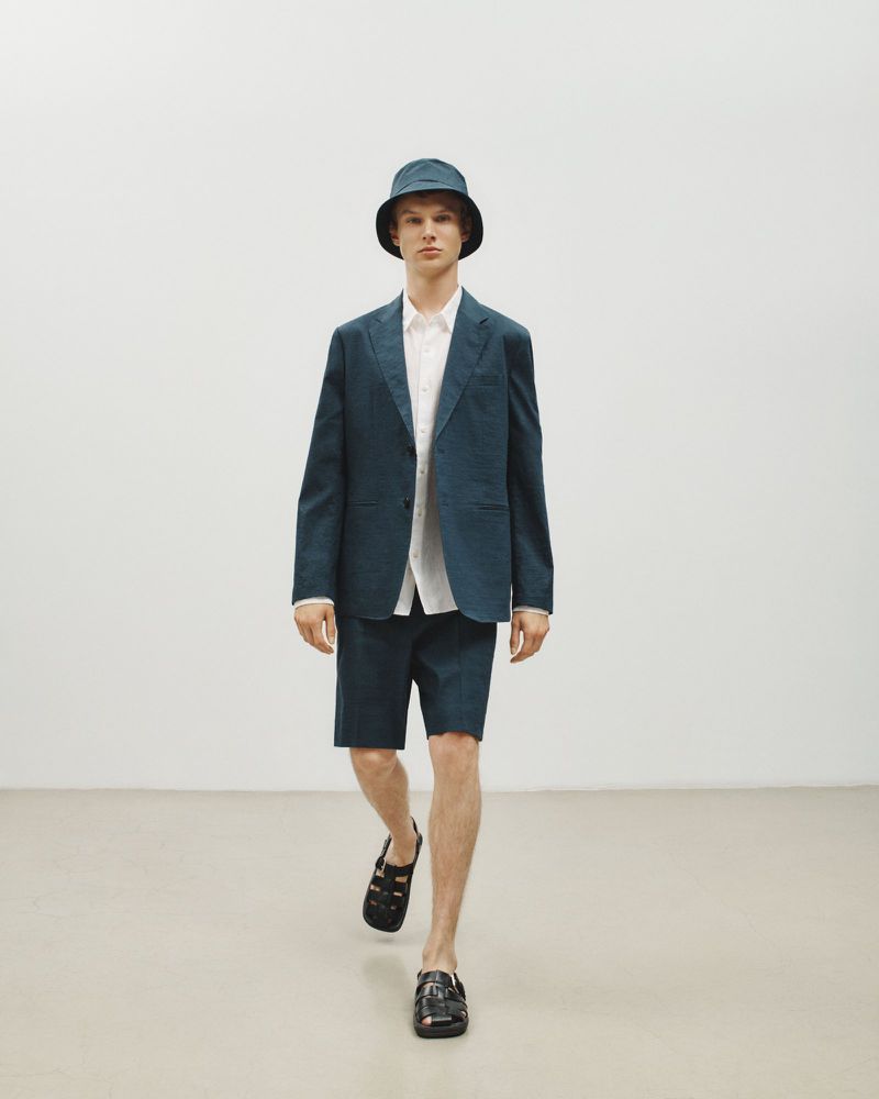 Model wearing dark blazer, matching shorts, bucket hat, and sandals walking forward on white studio seamless.