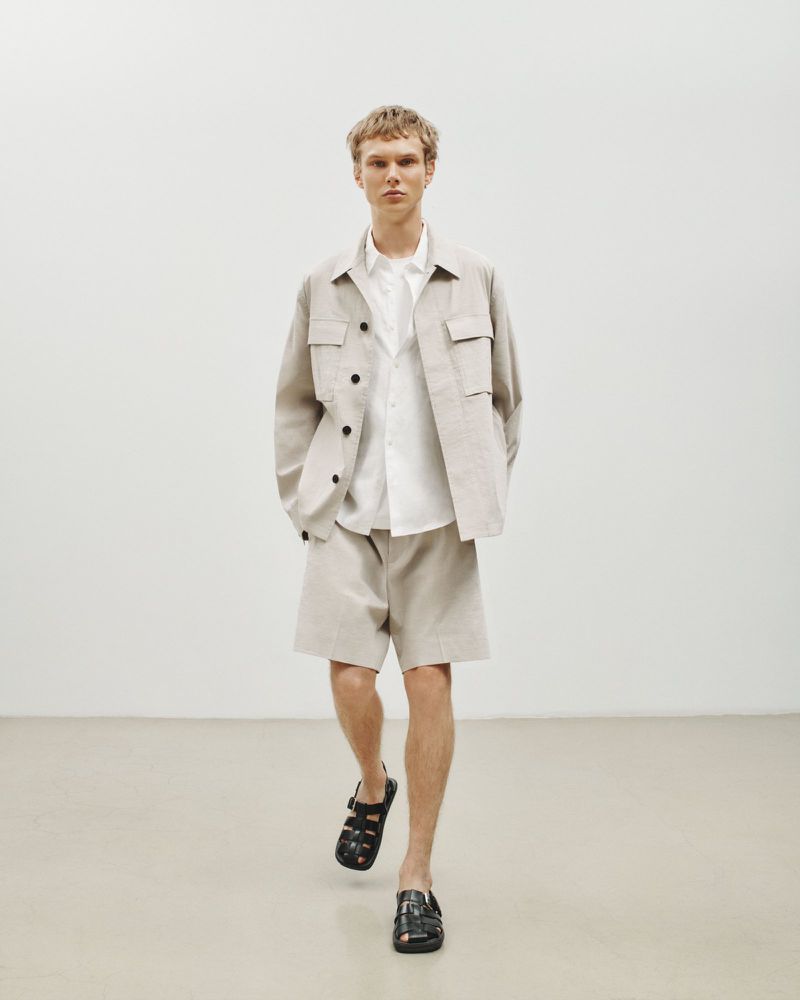 A fair-skinned male model poses in a light beige short suit with an open white button-down shirt, and black sandals in a studio setting.