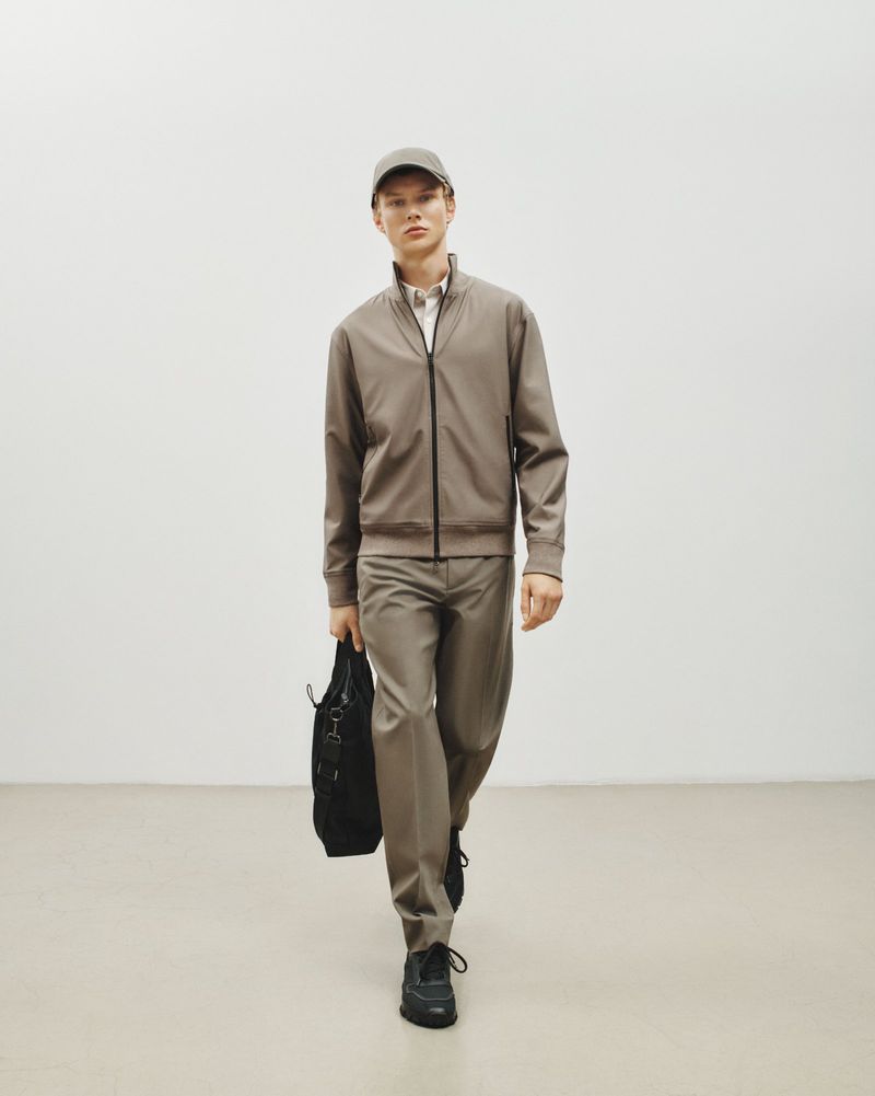 Front view of a male fashion model walking toward the camera wearing a jacket, dress pants, a baseball cap, and holding a black tote bag.