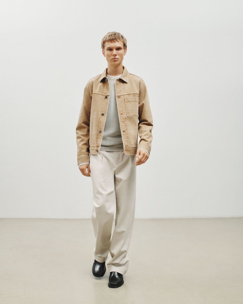 Man in beige jacket and white pants walks in minimalist studio. Fashion shot with soft light, showing modern style and simple background.