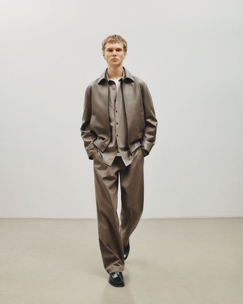 A young male model with short hair stands facing the camera wearing a brown collared leather jacket, button-down shirt, and loose trousers.