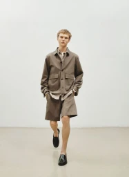 Male model wearing brown jacket and shorts walking toward camera against a plain white studio background