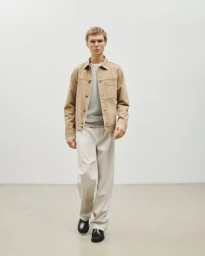 Full-length portrait of a male model wearing a tan denim jacket, light grey sweater, and white trousers against a neutral white background