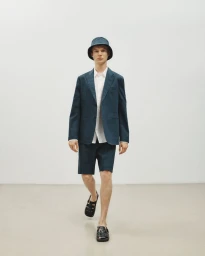 Model wearing dark blazer, matching shorts, bucket hat, and sandals walking forward on white studio seamless.