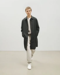 Model wearing a dark long coat over casual trousers walking against a plain studio background