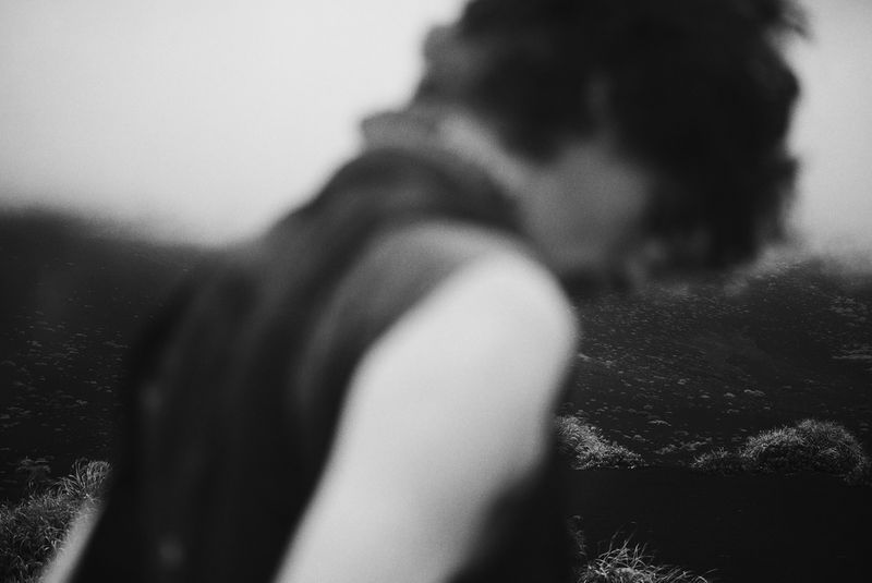 Monochrome blurred shot of a person with dark hair against a soft landscape with greenery. Focus is deliberately soft, creating an ethereal feel.
