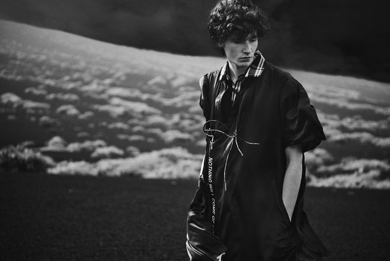 Monochrome portrait of a young adult male fashion model wearing an oversized top against a cloudy outdoor background on location.