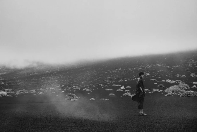 Monochrome portrait of a person standing on a volcanic landscape with sparse vegetation under a layer of low-lying fog.