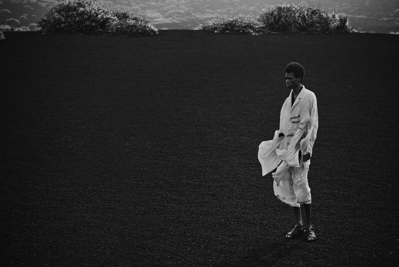 Black and white shot of a slender adult male in a windy field wearing a light-colored coat, gazing off in the distance. Cloudy sky.
