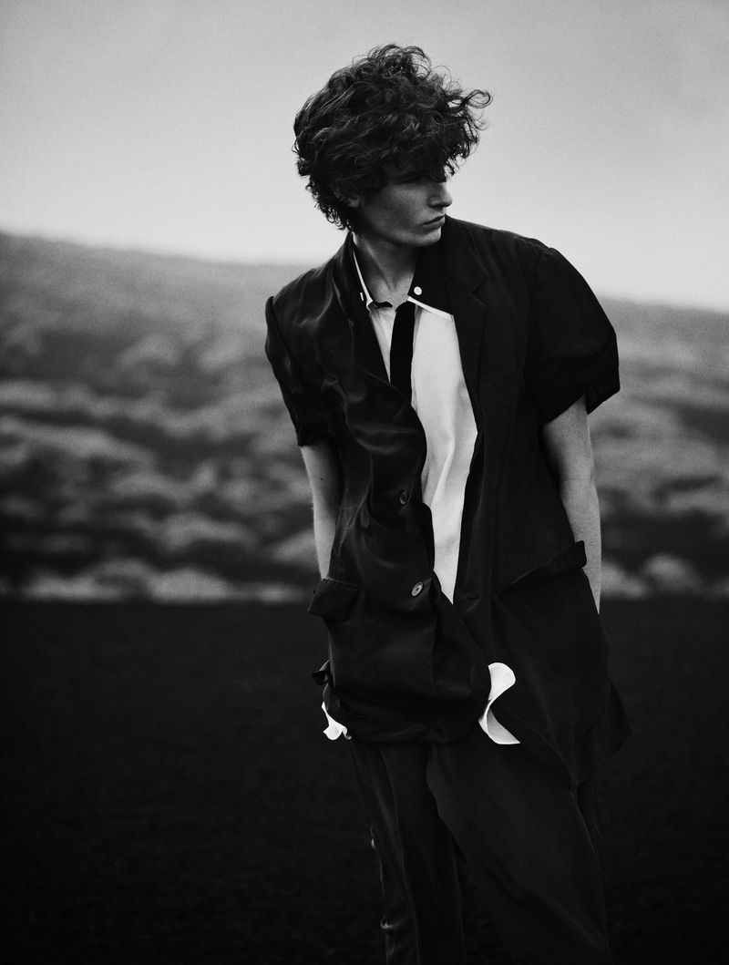 Black and white portrait of adult man outdoors with curly dark hair. The man is wearing a fashionable open shirt and pants.