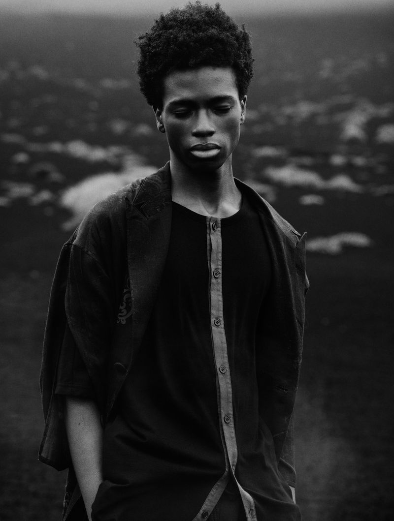 A young Black male model poses outdoors wearing a black button-down shirt against an out-of-focus landscape backdrop in monochrome.