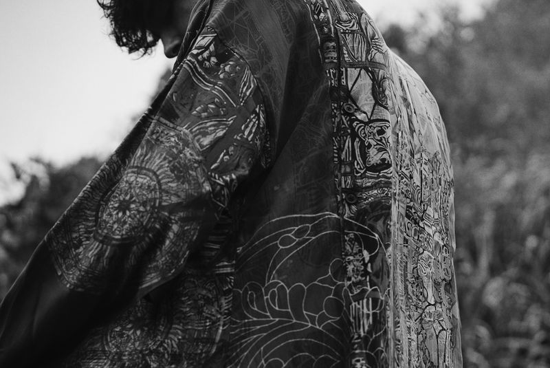 Black and white image of a person wearing a patterned kimono outside, with a blurred backdrop of trees and sky, showing only a hint of their face.