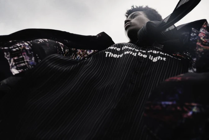 Low angle shot of a male model wearing a dark pinstripe shirt with white text against an overcast sky.