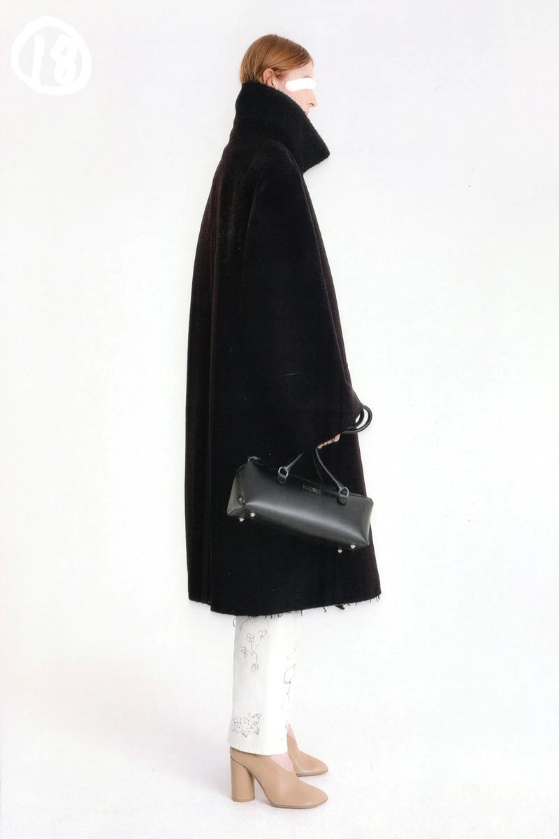 A woman in profile stands against a white background wearing a black coat and carrying a black bag with white print pants and nude heels.
