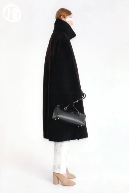 A woman in profile stands against a white background wearing a black coat and carrying a black bag with white print pants and nude heels.