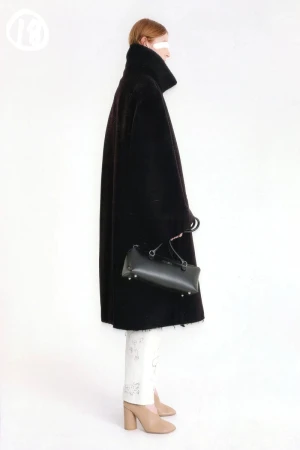 A woman in profile stands against a white background wearing a black coat and carrying a black bag with white print pants and nude heels.