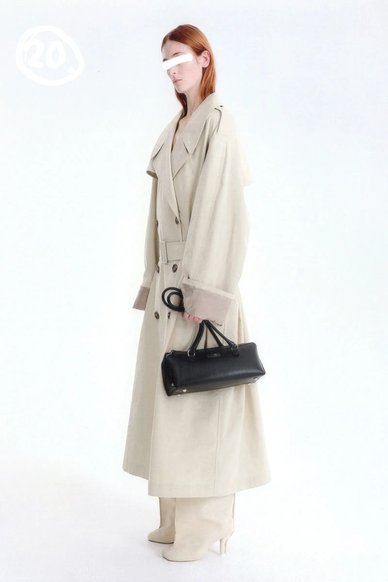 A person with auburn hair and an eye-mask standing in a long, beige double-breasted trench coat holding a black cylindrical handbag.