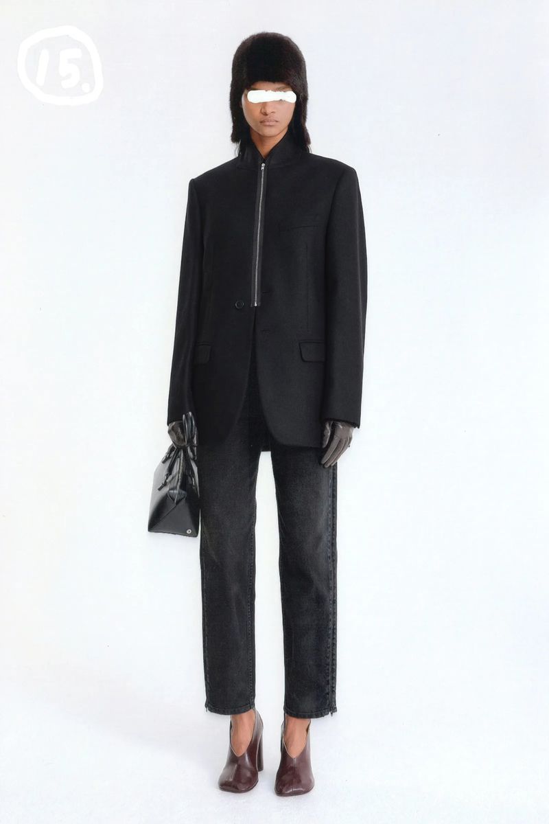 Full-length studio shot of a model wearing a black blazer, jeans, and heeled shoes while holding a black purse, with a white backdrop.