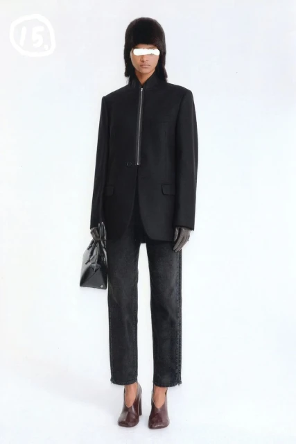 Full-length studio shot of a model wearing a black blazer, jeans, and heeled shoes while holding a black purse, with a white backdrop.