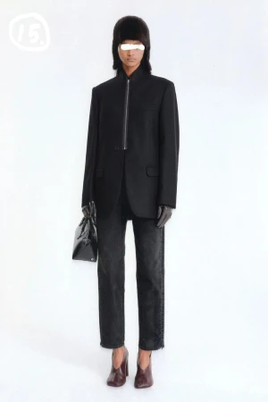Full-length studio shot of a model wearing a black blazer, jeans, and heeled shoes while holding a black purse, with a white backdrop.