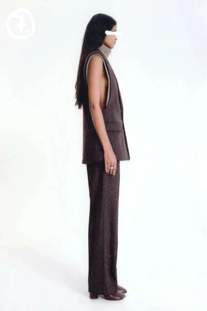 A woman in a brown sleeveless suit with long dark hair stands in a side profile pose against a bright white backdrop in a fashion editorial.