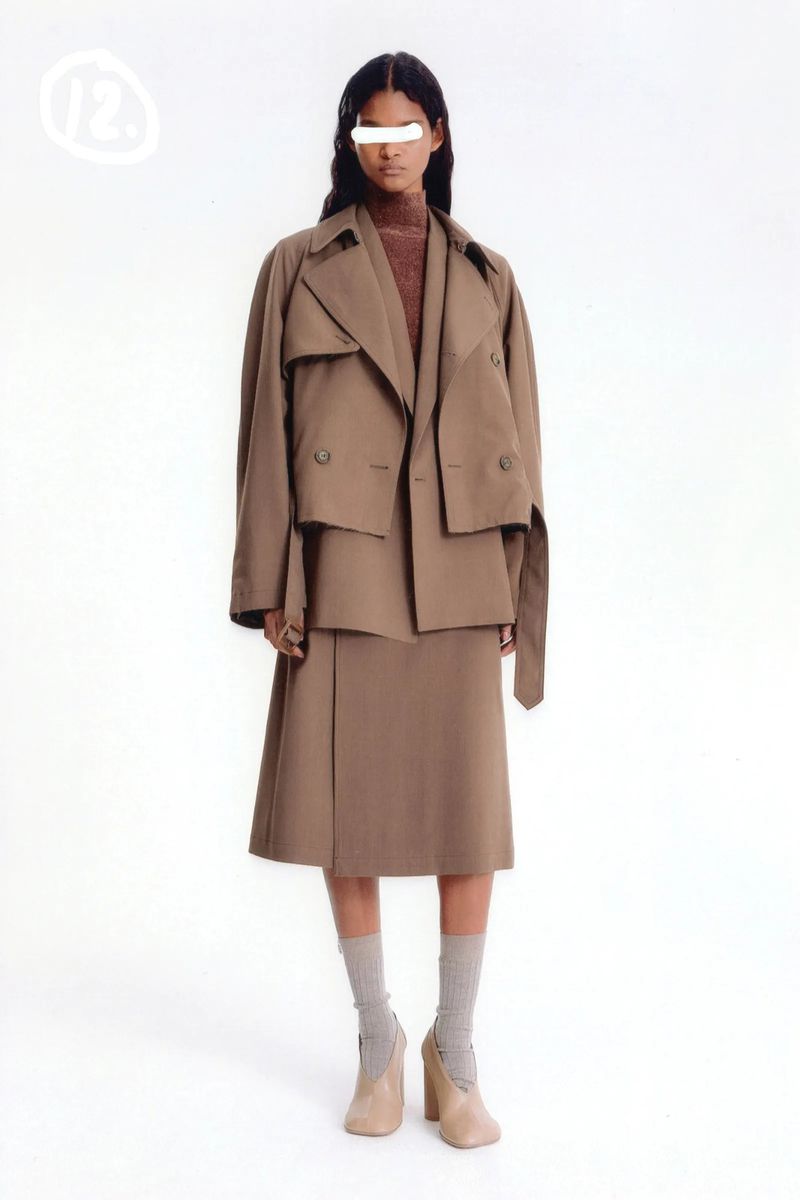 Woman in a brown trench coat and skirt, paired with gray socks and beige heels, poses against a bright white backdrop in a studio setting.
