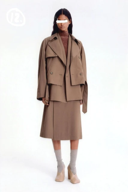 Woman in a brown trench coat and skirt, paired with gray socks and beige heels, poses against a bright white backdrop in a studio setting.