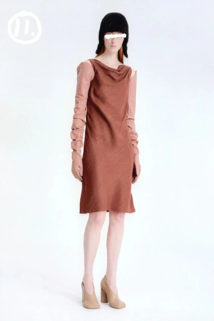A woman wears a draped-neck, knee-length dress with long sleeves, paired with heeled shoes and a cap against a clean white backdrop.