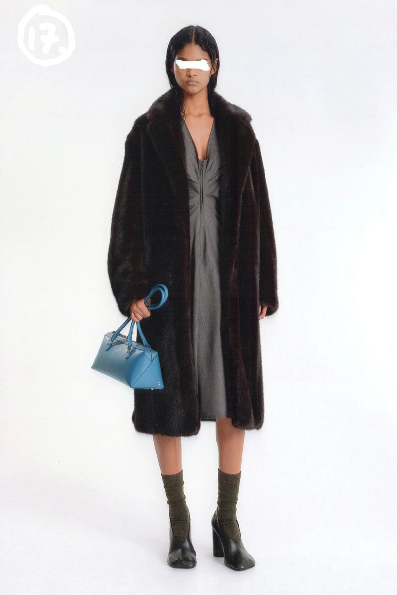 An adult woman stands in a photo studio wearing a grey dress and a brown faux fur coat, carrying a blue handbag and wearing black heeled boots.