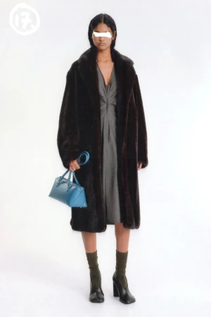 An adult woman stands in a photo studio wearing a grey dress and a brown faux fur coat, carrying a blue handbag and wearing black heeled boots.