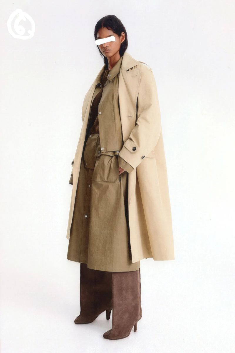 Adult female poses in studio wearing beige trench coat, tan shirt dress, and brown high-heeled boots in a fashion editorial shot.