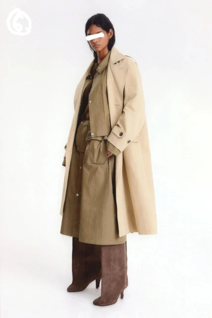 Adult female poses in studio wearing beige trench coat, tan shirt dress, and brown high-heeled boots in a fashion editorial shot.